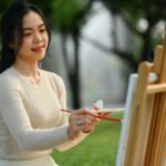 Smiling young woman painting a picture in the park. Art therapy and creative hobbies concept